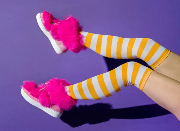 Side view of anonymous female with shadow of raised legs in yellow colored long socks and pink fuchsia shoes while sitting on violet background