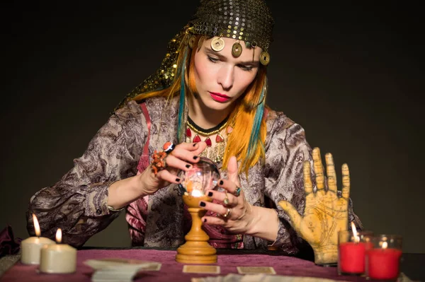 stock image Concentrated young female fortune teller with long ginger hair in dress and bandana sitting at table with burning candles cards and crystal ball while predicting destiny in dark room