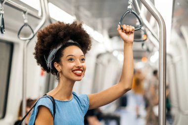 Afro saç bandı takmış, gülümseyen ve başını çeviren, yolculuk sırasında bulanık ve aydınlık metro treninde elinde tutarak duran Faslı bir kadın.