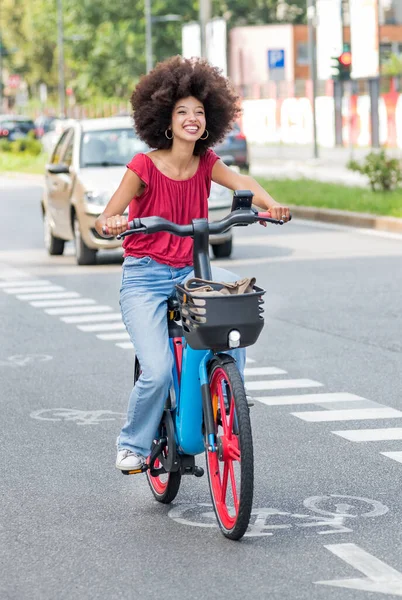 Afro saçlı olumlu genç Faslı kadın güneş ışığında bulanık binalara ve yeşil ağaçlara karşı asfalt yolda elektrikli bisiklet sürerken kafasını çeviriyor.