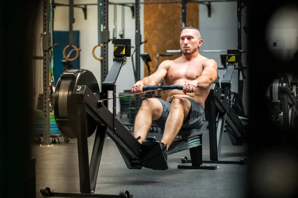 Full body of shirtless muscular male athlete in shorts training on rower machine during intense workout at fitness club