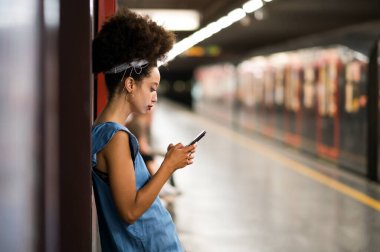 Gece metro istasyonunda cep telefonu ekranına bakıp mesajları kontrol eden Afro saçlı genç Faslı bir kadının yan görüntüsü.