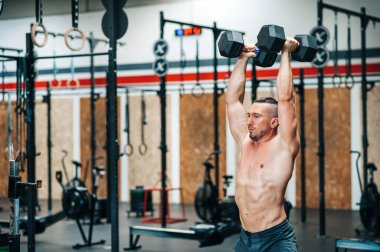 Modern ekipmanlarla bulanık spor salonunda dikilirken ve dambıllar üzerinde şeytan egzersizi yaparken odaklanmış genç, kaslı, üstsüz erkek atlet gözlerini kaçırıyor.