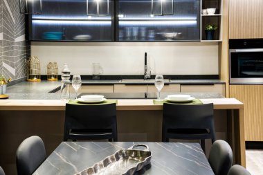 Mobilyalarla döşenmiş modern mutfağın içi mutfak rafları mutfak gereçleri sobanın yanındaki su şişesi şeffaf bardaklar yemek masası ve ışıklarla süslenmiş sandalyeler.