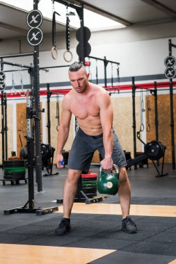 Spor salonunda ekipmanların yanında durup ağır kettlebell kapma egzersizine başlarken kaslı, çıplak erkek sporcu aşağıya bakıyor.