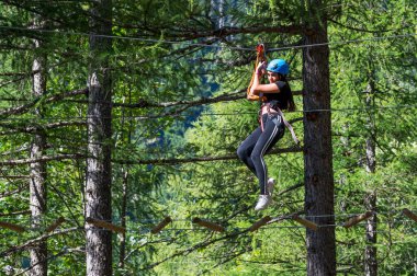 Kasklı heyecanlı genç bayanın düşük açılı görüntüsü güvenlik halatlarıyla ipte sallanırken ve gün ışığında orman macera parkında askıya alınmış teller üzerinde yürürken.