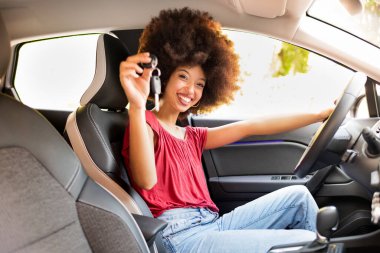 Afro saç stili mutlu etnik bayan arabada oturuyor ve yeni anahtar gösterirken kameraya bakıyor.