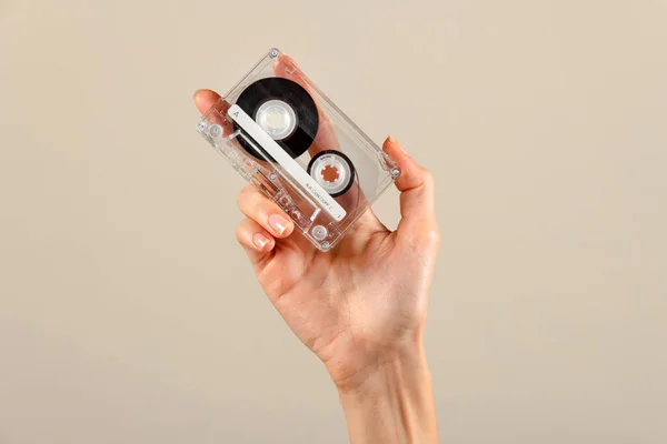 Anonymous female open palm hand with manicured reflecting nail fingers holding transparent retro cassette with black tape on wheels against beige background in light