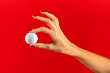 Elinde ojeli ince işaret ve başparmak parmaklarıyla golf topu tutan isimsiz genç bir kadın kırmızı arka planda kırmızı arka plana karşı gösteri yapıyor.