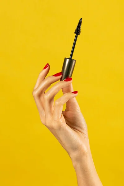 Anonymous young slim female with nails polished slender fingers holding aloft mascara base gently and demonstrating against yellow background in light