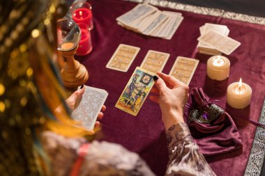 Elinde mumlar ve kristallerle masada otururken kostümlü, ölü tarot kartı seçen tanınmamış kadın falcının arka planda kalan görüntüsü.