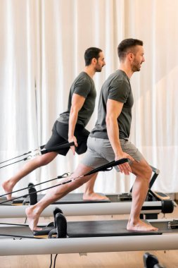 Spor kıyafetleri içindeki güçlü adamların beden eğitimi sırasında ıslah yataklarında direniş bantlarıyla pilates yapmaları.
