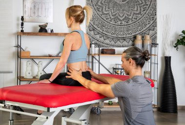 Modern klinikte tedavi sırasında arka planda duvar halısı ve rafları olan kırmızı sedye üzerinde oturan at kuyruklu kadın hastayı muayene eden odaklanmış fizyoterapistin yan görüntüsü