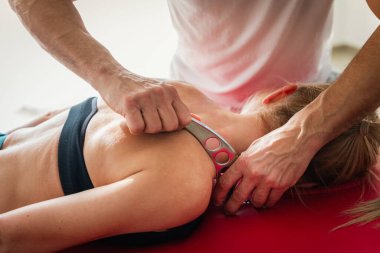 Crop massage therapist doing massage with IASTM tool on shoulder of female patient lying in wellness center during rehabilitation treatment at therapy room clipart