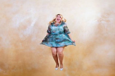 Ayrıca neşeli bir yüz ifadesi olan cüsseli bir kadın, sıcak bir stüdyo arkaplanının önünde, elbisesinin dalgalandığı bir ortamda yakalandı.