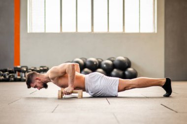 Sakallı, üstsüz, yetişkin bir erkeğin spor salonunda yerde yatıp, gün ışığında spor aletlerinin yanında şınav çekerken yan görünüşü.