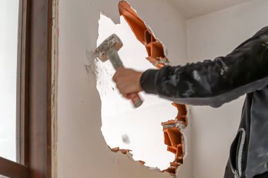 Construction worker using sledgehammer to demolish interior brick wall, creating hole during home renovation clipart