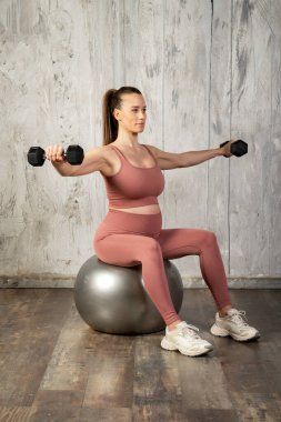 Hamile bir kadın spor topunun üzerinde otururken dambıllarıyla yan yana yükselerek sağlıklı hamileliği ve aktif yaşam tarzını teşvik ediyor.