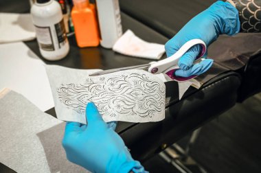 Dövme sanatçısı mavi eldiven takıyor şablonu makasla kesiyor, dövme oturumu için hazırlıyor.