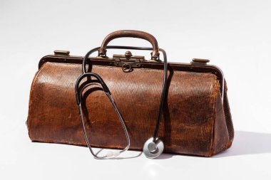 Beyaz arka planda steteskoplu eski kahverengi deri doktor çantası, sağlık ve tıbbi geçmişi temsil ediyor.