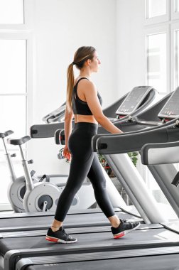 Parlak bir spor salonunda koşu bandında yürürken formda bir kadın ısınıyor.