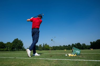 Golf sopasını golf sahasında sallıyor, açık mavi gökyüzüne doğru bir golf topuna vuruyor.