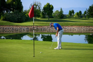 Kıdemli golfçü güneşli bir günde su tehlikesinin yanında güzel bir yeşil giysi giyiyor.