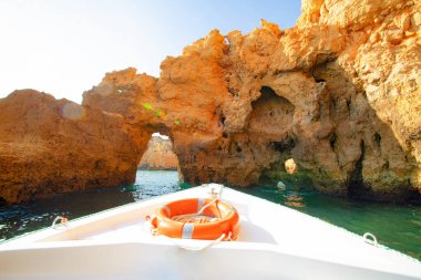 Güzel kayalıkların ve kemerlerin arka planında turuncu can simidi olan beyaz bir tekne.