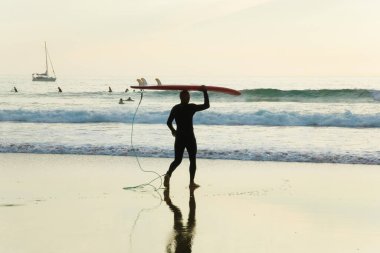 Arrifana, Aljezur, Portekiz. 09 numara. 29 yaşında. 2020. Gün batımı plajında sörf tahtası taşıyan sörfçü. Sörfçü spor sağlığı ve seyahat konsepti.