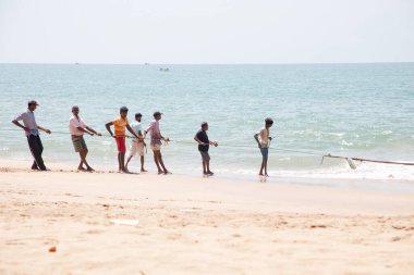 Unawatuna, Sri Lanka 09 Şubat 2023 'te farklı renklerde yaz kıyafetleri giyen bir grup adam balıkçıların Güney Sri Lanka sahilindeki büyük balık ağını çekmelerine yardım ediyor. Sıkı takım çalışması..