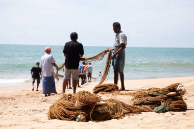 Unawatuna, Sri Lanka 09 Şubat 2023 'te farklı renklerde yaz kıyafetleri giyen bir grup adam balıkçıların Güney Sri Lanka sahilindeki büyük balık ağını çekmelerine yardım ediyor. Sıkı takım çalışması..