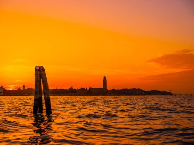 Venedik, İtalya. 29 Ekim 2022 Venedik silueti gün batımında batan güneşin parlak turuncu-kırmızı ışınlarında. Sudan manzara. Geleneksel Venedik kümeleri sudan sarkan