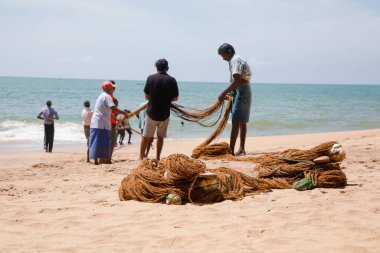 Unawatuna, Sri Lanka 09 Şubat 2023 'te farklı renklerde yaz kıyafetleri giyen bir grup adam balıkçıların Güney Sri Lanka sahilindeki büyük balık ağını çekmelerine yardım ediyor. Sıkı takım çalışması..