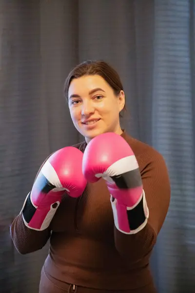 a young woman in boxing gloves on a gray background looks at the camera, stands in a boxing stance
