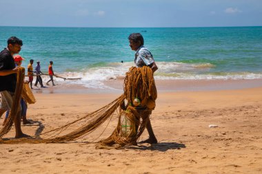 Unawatuna, Sri Lanka 09 Şubat 2023 'te farklı renklerde yaz kıyafetleri giyen bir grup adam balıkçıların Güney Sri Lanka sahilindeki büyük balık ağını çekmelerine yardım ediyor. Sıkı takım çalışması..