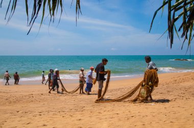 Unawatuna, Sri Lanka 09 Şubat 2023 'te farklı renklerde yaz kıyafetleri giyen bir grup adam balıkçıların Güney Sri Lanka sahilindeki büyük balık ağını çekmelerine yardım ediyor. Sıkı takım çalışması..