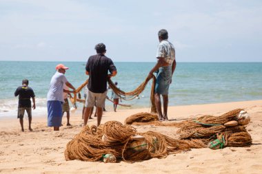 Unawatuna, Sri Lanka 09 Şubat 2023 'te farklı renklerde yaz kıyafetleri giyen bir grup adam balıkçıların Güney Sri Lanka sahilindeki büyük balık ağını çekmelerine yardım ediyor. Sıkı takım çalışması..