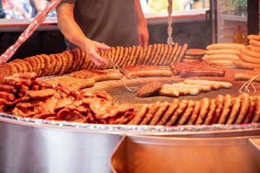 Farklı türde sosis, köfte ve pişirilen etle dolu büyük ızgara. Birisi ızgara yapıyor, sosis hazırlamak için maşa kullanıyor. Sahne bir gıda pazarı ya da festivalde gibi görünüyor..