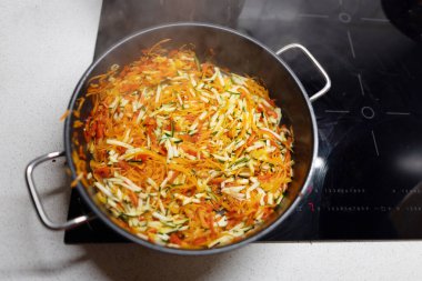 İnce doğranmış renkli mevsimlik sebzelerle büyük yuvarlak, modern bir tava pişirmeye hazır endüksiyon yüzeyinde duruyor.