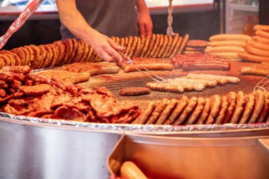 Farklı türde sosis, köfte ve pişirilen etle dolu büyük ızgara. Birisi ızgara yapıyor, sosis hazırlamak için maşa kullanıyor. Sahne bir gıda pazarı ya da festivalde gibi görünüyor..