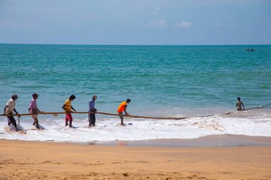 Unawatuna, Sri Lanka 09 Şubat 2023 'te farklı renklerde yaz kıyafetleri giyen bir grup adam balıkçıların Güney Sri Lanka sahilindeki büyük balık ağını çekmelerine yardım ediyor. Sıkı takım çalışması..