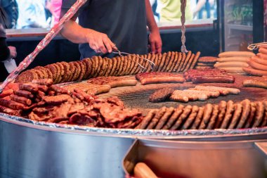 Farklı türde sosis, köfte ve pişirilen etle dolu büyük ızgara. Birisi ızgara yapıyor, sosis hazırlamak için maşa kullanıyor. Sahne bir gıda pazarı ya da festivalde gibi görünüyor..