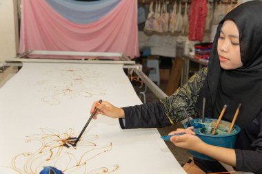 Kuala Lumpur, Malezya. 04.08.2024. Türbanlı genç bir Müslüman kız fırçayla kumaşa geleneksel desenler çiziyor. Kuala lumpur 'daki Batik fabrikası.