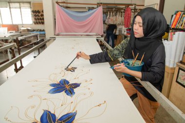 Kuala Lumpur, Malezya. 04.08.2024. Türbanlı genç bir Müslüman kız fırçayla kumaşa geleneksel desenler çiziyor. Kuala lumpur 'daki Batik fabrikası.