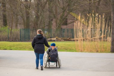 Berlin, Almanya 05.03.2024. Parkta tekerlekli sandalyede oturan bir çocukla yürüyen bir kadın.