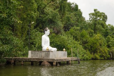 Beruwala,Sri Lanka. 07 Şubat 2023. Beyaz Buda figürü gölün üzerinde oturuyor. Arka plan güzel doğa ağaçları, mavi gökyüzü. Asya seyahati ve din kavramı