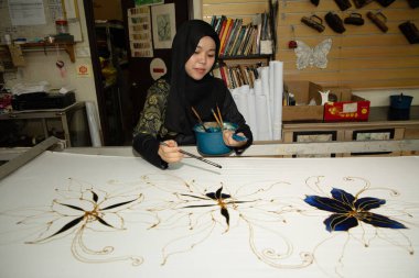 Kuala Lumpur, Malezya. 04.08.2024. Türbanlı genç bir Müslüman kız fırçayla kumaşa geleneksel desenler çiziyor. Kuala lumpur 'daki Batik fabrikası.