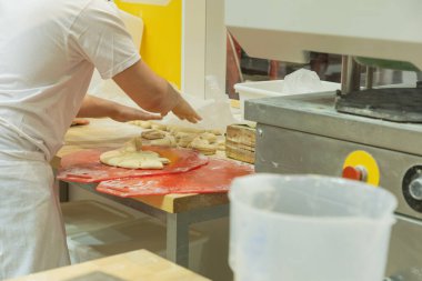 Profesyonel fırıncı, çeşitli pişirme aletleri ve malzemeleriyle çevrili yoğun bir mutfakta kırmızı kesme tahtasında hamur şekillendiriyor..