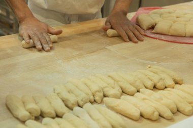 Baker ahşap bir tezgahta hamuru şekillendiriyor, yeni oluşmuş ekmek ruloları ile çevrili, sıcak bir mutfak atmosferi yaratıyor..