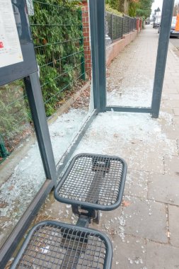 Kırık cam ve molozların bulunduğu hasarlı otobüs durağı barınağı yeşillik ve kentsel çevre ile çevrili..